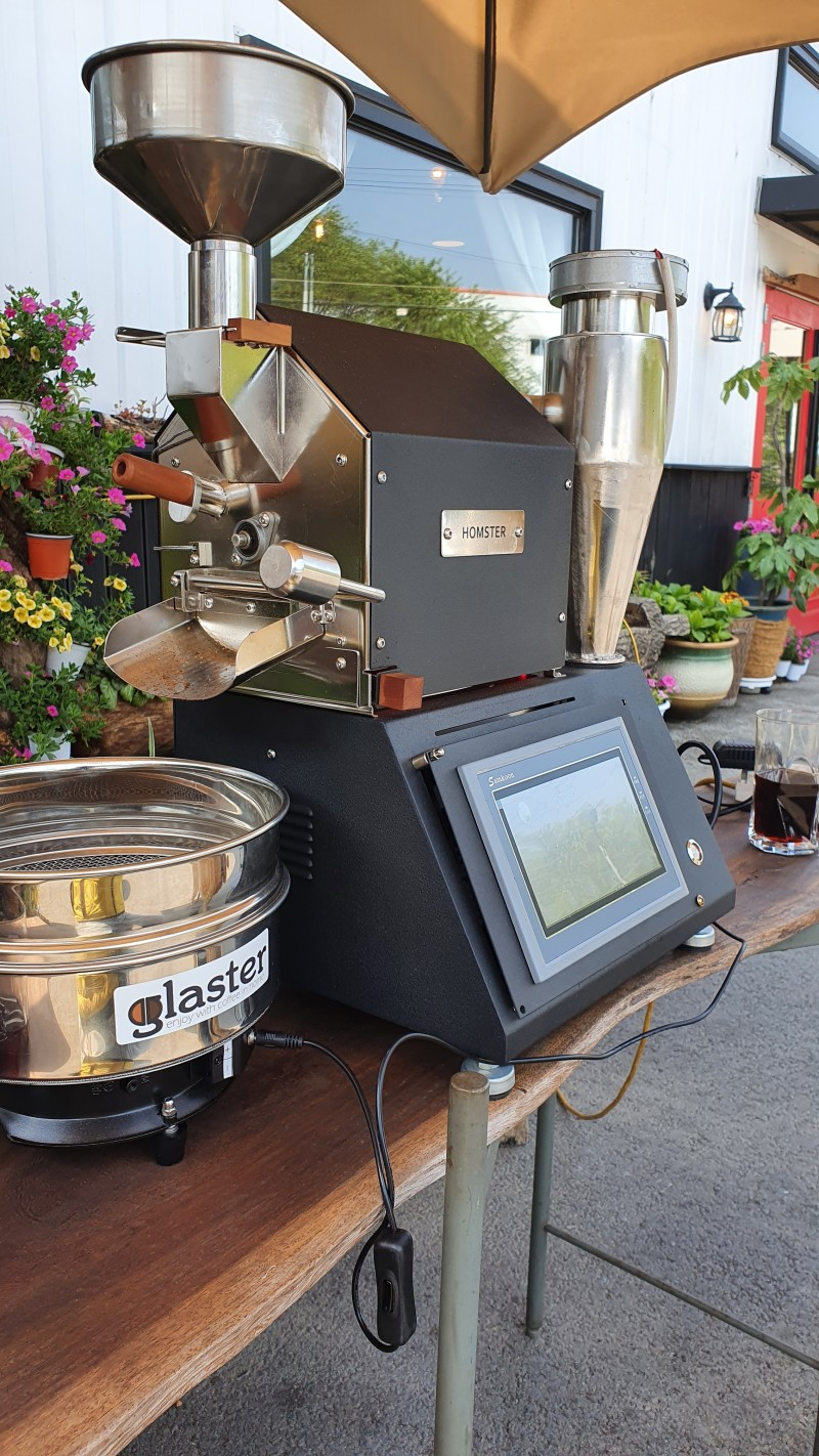 Coffee My Cup: Coffee roasting machine on a wooden table outdoors with plants and a building in the background