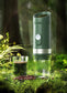 Coffee My Cup: Green portable coffee maker on a grassy background with a glass of coffee and coffee beans.